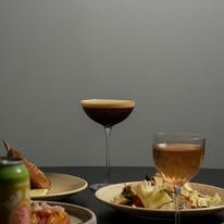 Espresso martini coupe alongside dinner plates on a dark table with grey background